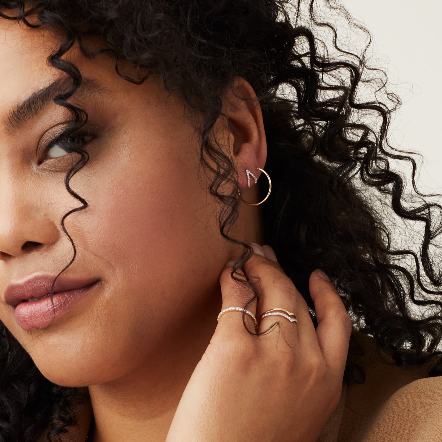 Woman with curly hair wearing a PavÃ© Diamond Wishbone Ring and gold earrings, close-up on ring and earrings highlighting the intricate design and craftsmanship.