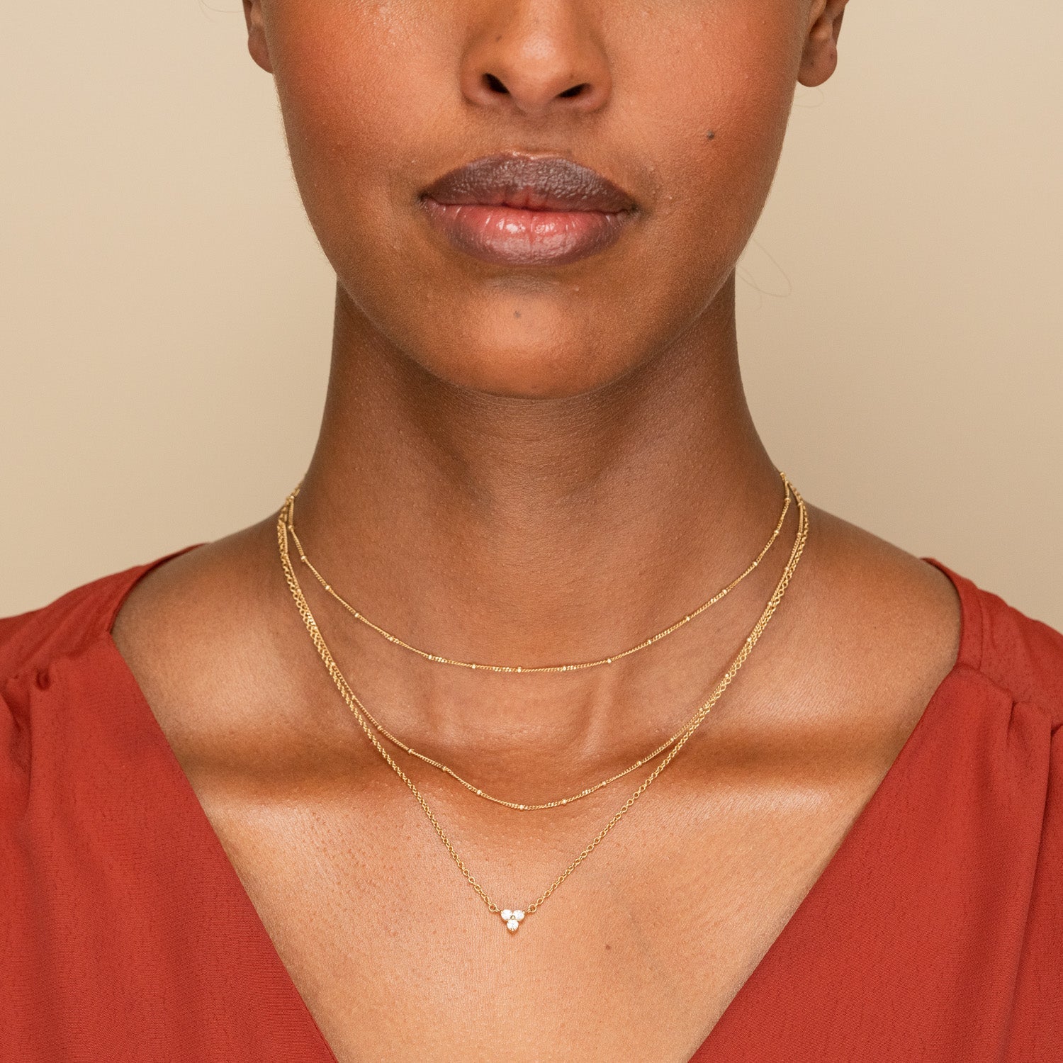 Woman wearing the versatile Lotus Necklace, featuring three fine gold chains with a triangular pendant of tiny stones, elegantly layered over a rust-red v-neck top.