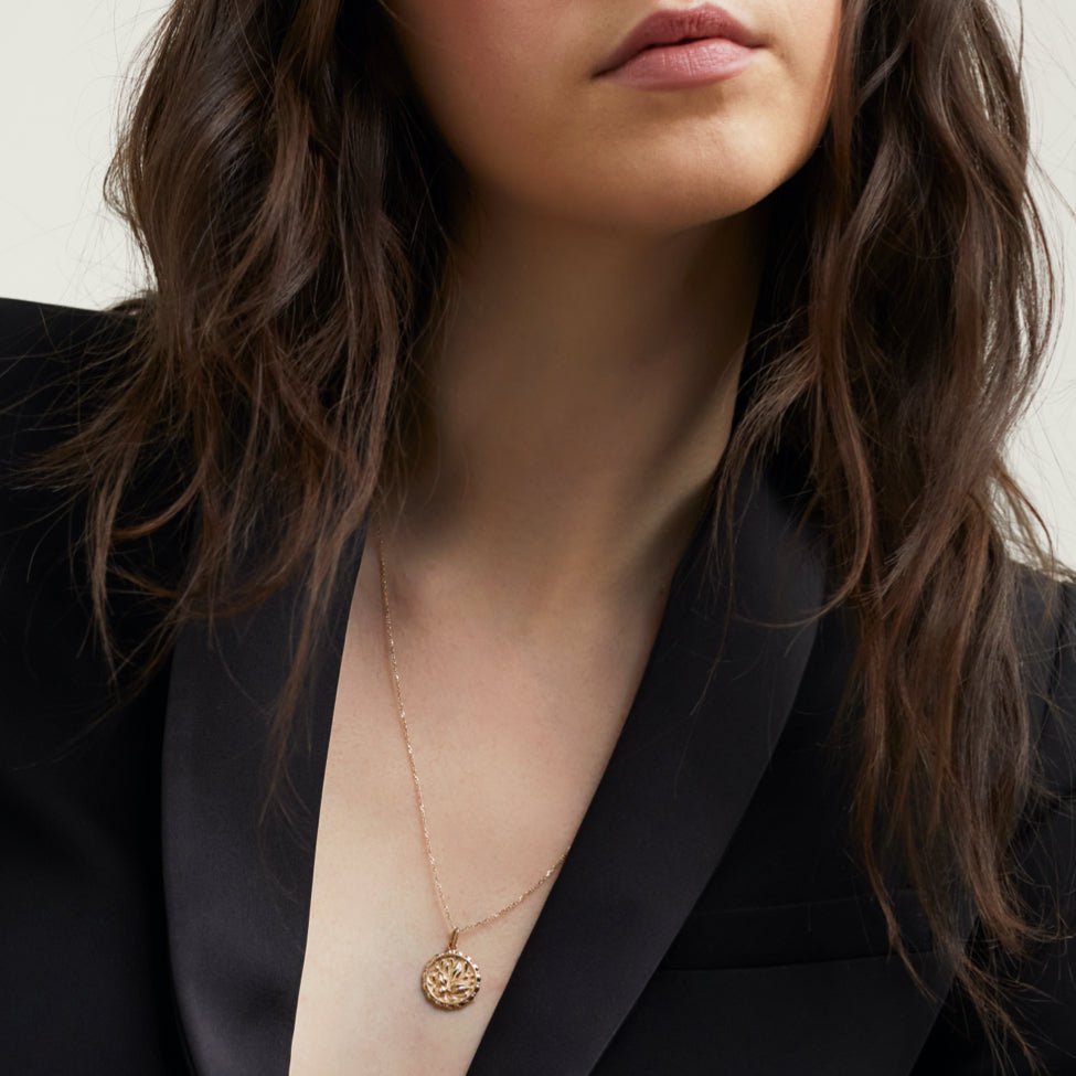 Woman wearing a black blazer, showcasing the Balance: Terra Coin Charm Pendant, a gold medallion inspired by Greek Goddess Gaia, symbolizing individual balance amidst chaos.