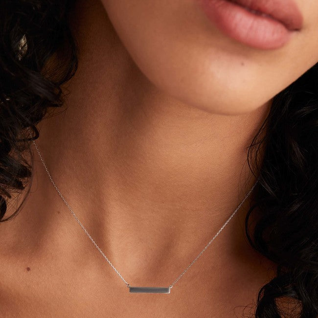 A woman wearing the Horizontal Engravable Bar Necklace, handcrafted in 14k solid gold with a sleek, minimal design, complemented by earrings and lipstick.